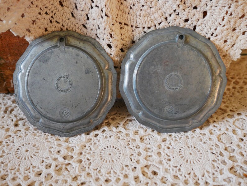 May include: Two silver-toned metal plates with scalloped edges. The back of each plate has a circular design with the text "Atelier d'Art" and "Petites Maisons" in a circle.