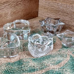 May include: A collection of clear glass candle holders in various shapes: two hearts, a star, and a five-pointed star. The candle holders are on a textured surface with a wooden background. The glass is transparent, allowing light to pass through.