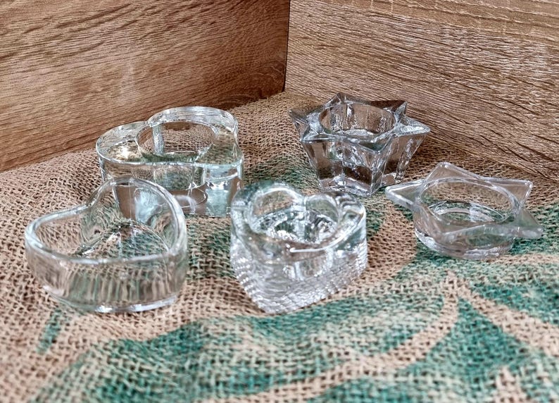 May include: A collection of clear glass candle holders in various shapes: two hearts, a star, and a five-pointed star. The candle holders are on a textured surface with a wooden background. The glass is transparent, allowing light to pass through.
