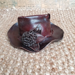 May include: A brown leather hat with a flower design. The hat has a wide brim and a tall crown. The flower is made of leather and is dark brown.