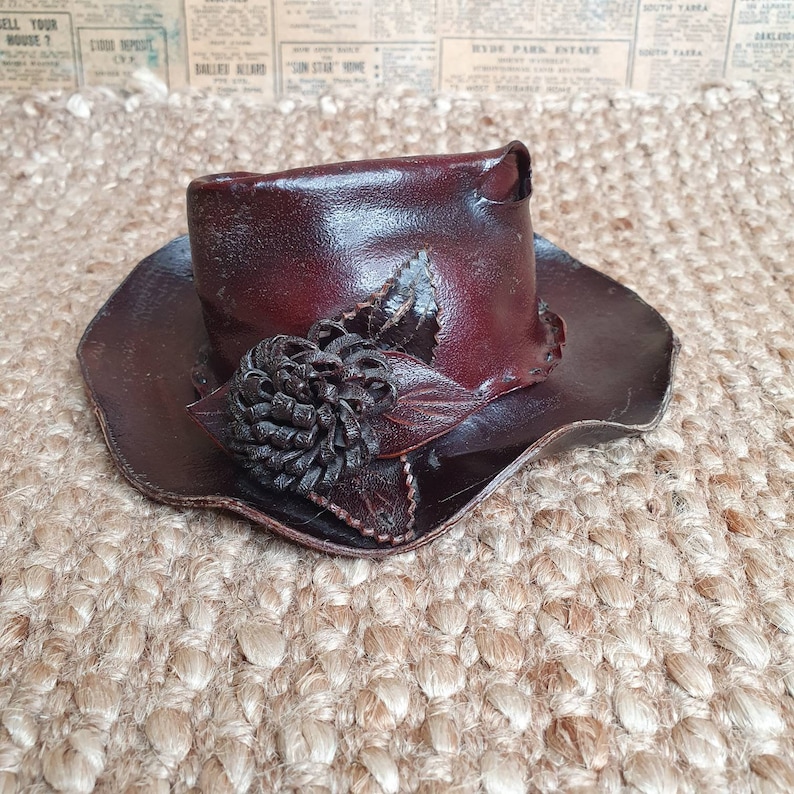 May include: A brown leather hat with a flower design. The hat has a wide brim and a tall crown. The flower is made of leather and is dark brown.