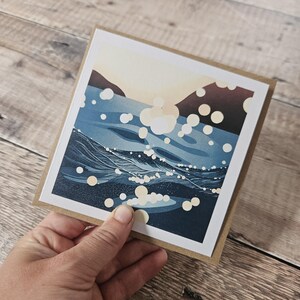 Pu&ograve; includere: Biglietto di auguri quadrato con una scena oceanica blu e bianca con un tramonto e bolle bianche. Il biglietto &egrave; tenuto in mano e ha un bordo marrone.