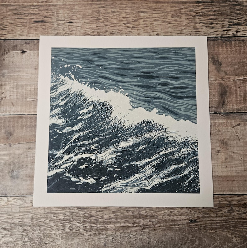 May include: A square print depicting a stylized ocean wave. The artwork features shades of blue and white, with the wave cresting and breaking. The print is set against a white border.