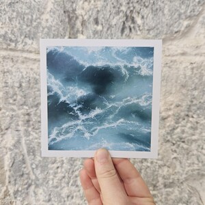 May include: A square print featuring an abstract ocean wave design in shades of blue and white. The image captures the movement and texture of water, with swirling patterns and foamy white crests. The print is held against a textured gray brick wall.