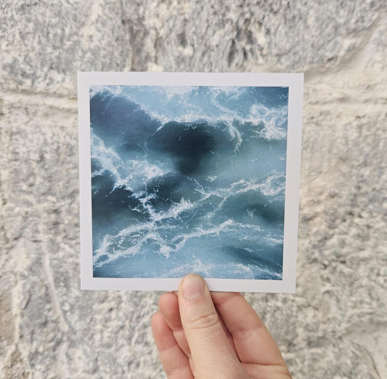 May include: A square print featuring an abstract ocean wave design in shades of blue and white. The image captures the movement and texture of water, with swirling patterns and foamy white crests. The print is held against a textured gray brick wall.