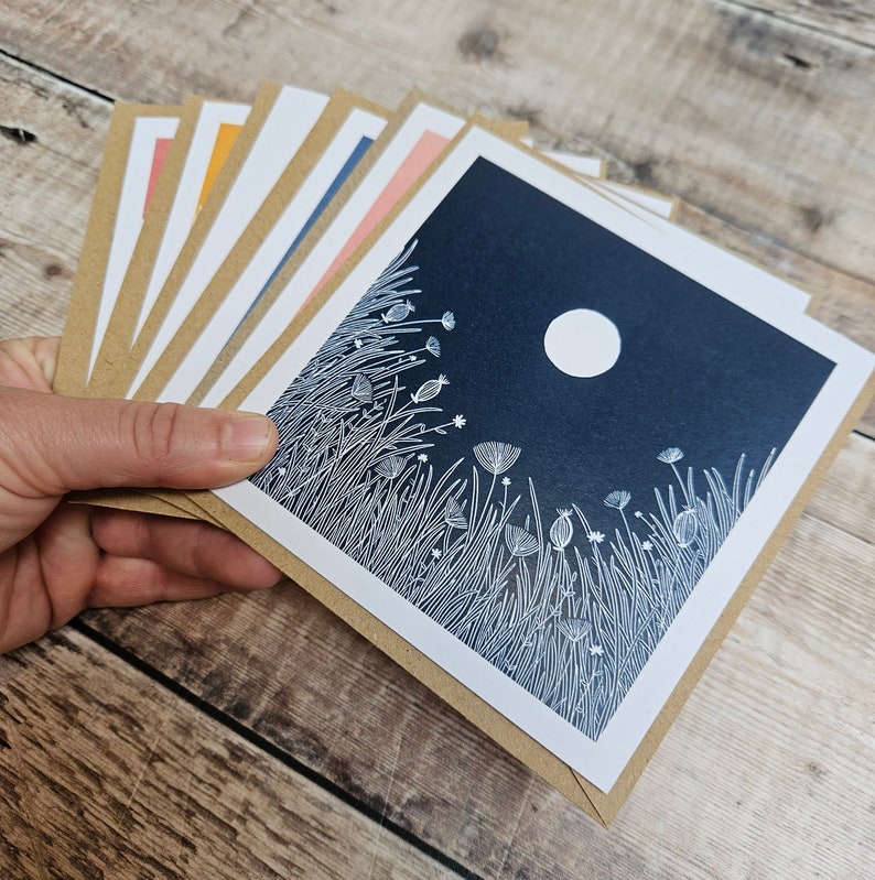 K&ouml;nnte beinhalten: Ein Satz von acht leeren Gru&szlig;karten mit braunen Umschl&auml;gen. Die Karten zeigen eine wei&szlig;e Linienzeichnung eines Mondes und Grasses mit Blumen auf dunkelblauem Hintergrund.