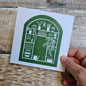K&ouml;nnte beinhalten: Eine gr&uuml;ne und wei&szlig;e Illustration eines Gew&auml;chshauses mit verschiedenen Pflanzen und einer Gie&szlig;kanne. Das Gew&auml;chshaus ist mit einem abgerundeten Bogen und Regalen voller Topfpflanzen dargestellt.