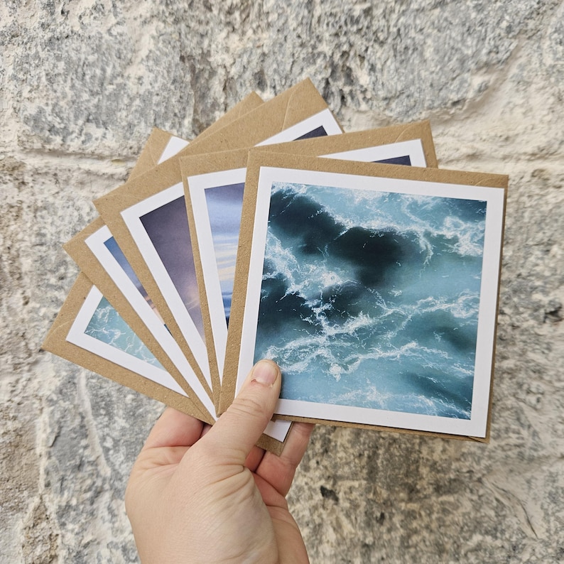 May include: A hand holds a fan of square greeting cards with ocean wave imagery. The cards have white borders and are set against brown kraft paper envelopes. The ocean images feature shades of blue and white, capturing the movement and texture of the water.