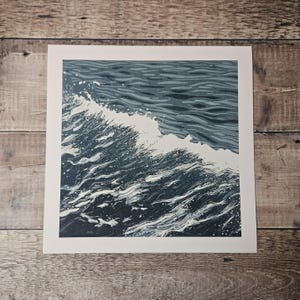 May include: A square print featuring a stylized depiction of ocean waves. The artwork uses shades of blue and white to create a dynamic image of a breaking wave. The print is set against a white border.
