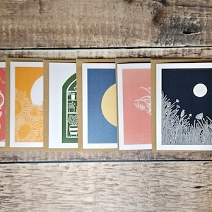 K&ouml;nnte beinhalten: Satz von sieben leeren Gru&szlig;karten mit verschiedenen Illustrationen. Die Karten zeigen eine Vielzahl von Designs, darunter einen Mond, Blumen, eine Teekanne, ein Fenster mit Pflanzen und eine Sonnenblume. Die Karten sind alle wei&szlig; mit schwarzen Umrissen und auf cremefarbenem Karton gedruckt.