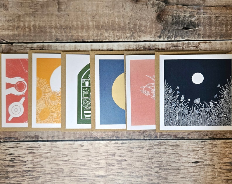 K&ouml;nnte beinhalten: Satz von sieben leeren Gru&szlig;karten mit verschiedenen Illustrationen. Die Karten zeigen eine Vielzahl von Designs, darunter einen Mond, Blumen, eine Teekanne, ein Fenster mit Pflanzen und eine Sonnenblume. Die Karten sind alle wei&szlig; mit schwarzen Umrissen und auf cremefarbenem Karton gedruckt.