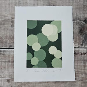 May include: A print with a dark green backdrop and overlapping circles in various shades of green and off-white. The artwork is titled "Green Bokeh" and is numbered 3/8. The print is on white paper with a deckled edge.
