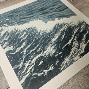 May include: A print depicting a stylized ocean wave. The artwork features shades of blue and white, with the wave cresting and breaking. The image has a textured, artistic quality, suggesting a linocut or woodcut style.