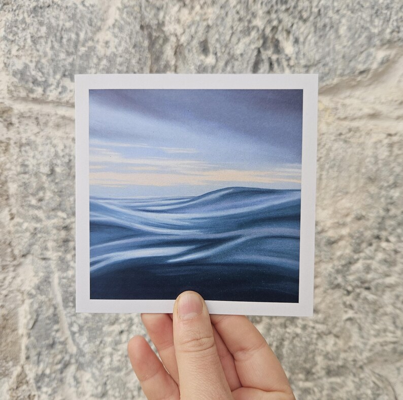 May include: A square art print featuring a serene ocean scene. The artwork depicts rolling blue waves under a cloudy sky with hints of pale yellow. The print is held against a textured gray stone wall.