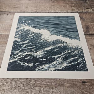 May include: A print of a wave in shades of blue and white. The artwork depicts the cresting wave with detailed lines and textures, capturing the movement and energy of the ocean. The print is on a white rectangular paper.