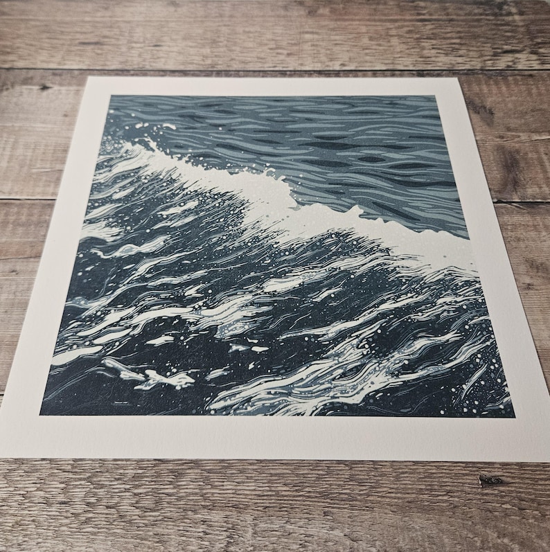 May include: A print of a wave in shades of blue and white. The artwork depicts the cresting wave with detailed lines and textures, capturing the movement and energy of the ocean. The print is on a white rectangular paper.