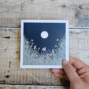 K&ouml;nnte beinhalten: Eine wei&szlig;e Linienzeichnung eines Feldes mit Blumen und Gras unter einem Vollmond vor einem dunkelblauen Hintergrund.