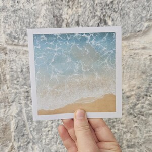 May include: A square print featuring an aerial view of a beach. The image shows the ocean's waves meeting the sandy shore. The water transitions from light blue to white as it approaches the beach. The print is held against a gray stone wall.