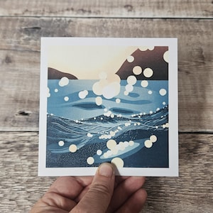 Pu&ograve; includere: Stampa artistica quadrata raffigurante un paesaggio marino con un orizzonte soleggiato e acqua blu scuro. Le onde sono rese con linee strutturate e il riflesso del sole &egrave; rappresentato da forme circolari bianche. La stampa &egrave; tenuta da una mano.