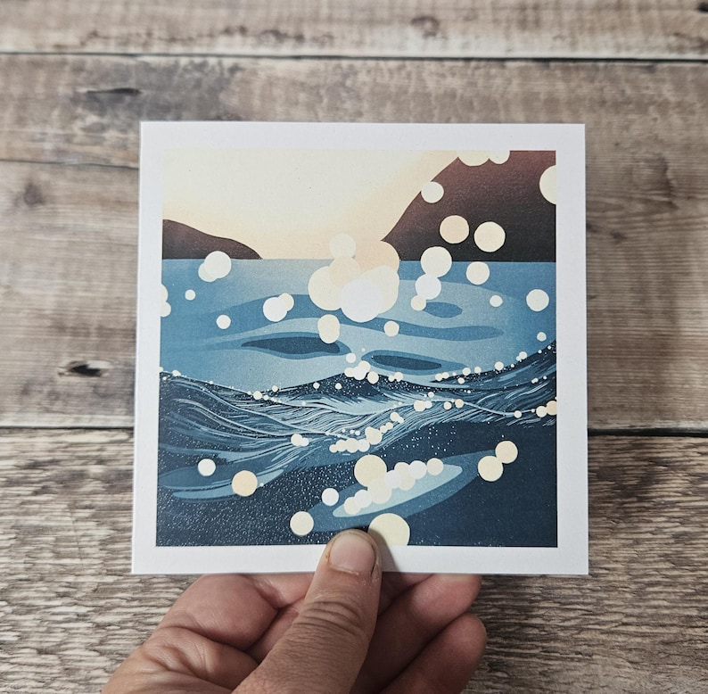 Pu&ograve; includere: Stampa artistica quadrata raffigurante un paesaggio marino con un orizzonte soleggiato e acqua blu scuro. Le onde sono rese con linee strutturate e il riflesso del sole &egrave; rappresentato da forme circolari bianche. La stampa &egrave; tenuta da una mano.