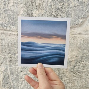 May include: A square art print featuring a serene ocean scene. The artwork depicts rolling blue waves under a dusky sky with hints of orange and gray. The print is held by a hand against a textured stone wall.