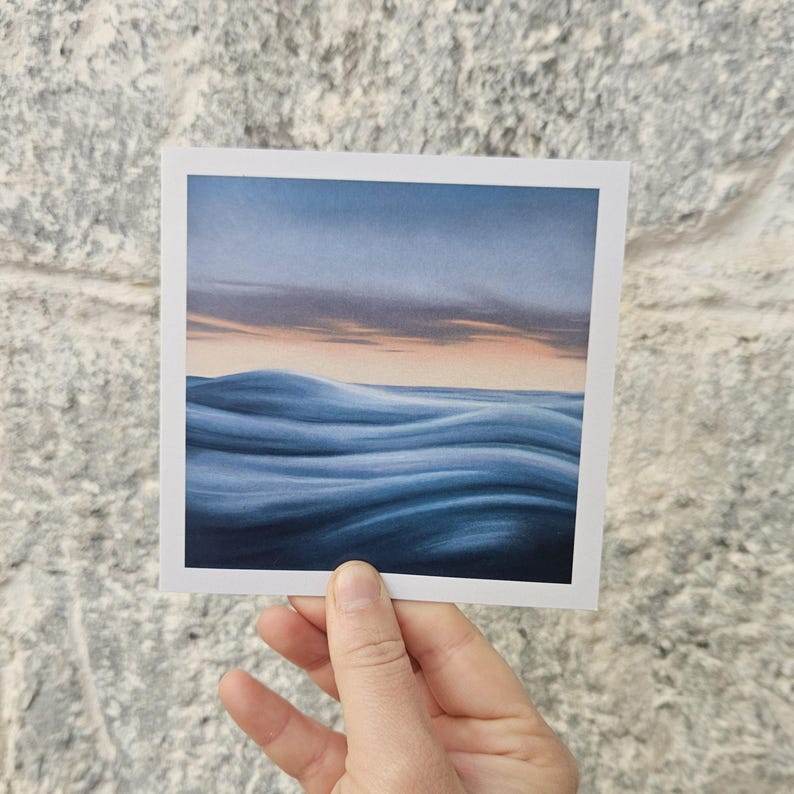May include: A square art print featuring a serene ocean scene. The artwork depicts rolling blue waves under a dusky sky with hints of orange and gray. The print is held by a hand against a textured stone wall.