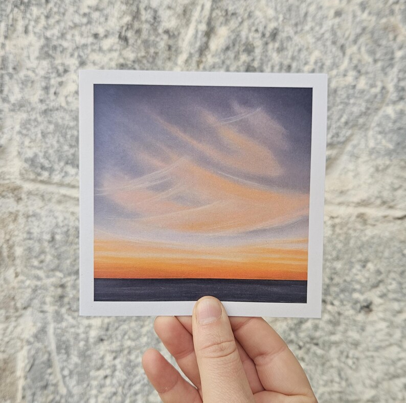 May include: A square art print featuring a sunset over the ocean. The sky transitions from light blue to orange, with wispy clouds. The ocean is a dark blue. The print is held by a hand against a textured gray brick wall.