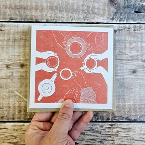 K&ouml;nnte beinhalten: Eine rote Karte mit einer wei&szlig;en Illustration von vier H&auml;nden, die Tassen mit Kaffee oder Tee halten. Die H&auml;nde reichen in Richtung der Mitte der Karte, wo sich auch eine Teekanne befindet.