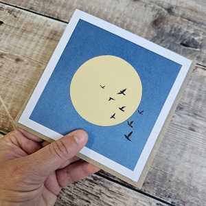 K&ouml;nnte beinhalten: Eine blaue Karte mit einem gelben Kreis und schwarzen Silhouetten von V&ouml;geln, die in V-Formation fliegen. Die Karte hat einen wei&szlig;en Rand.