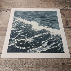 May include: A print of a wave breaking in the ocean. The artwork features shades of blue and white, with the wave cresting and creating a splash. The print is on a white background.