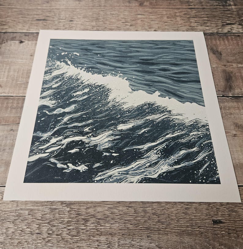 May include: A print of a wave breaking in the ocean. The artwork features shades of blue and white, with the wave cresting and creating a splash. The print is on a white background.