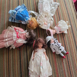 May include: Five small dolls arranged in a circle. One wears a blue and white dress, one a white lace gown, one a red and white striped dress, one a white dress with a pink sash, and one a white coat with a floral dress and a white hat.