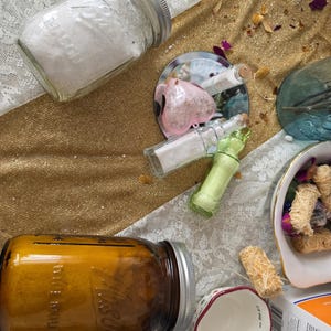 Könnte beinhalten: Eine Sammlung von Apothekergläsern und kleinen Flaschen, die mit verschiedenen Substanzen gefüllt sind. Die Gläser sind klar und bernsteinfarben, mit silbernen Deckeln. Eine herzförmige Flasche und ein kleiner Messbecher sind ebenfalls vorhanden, zusammen mit getrockneten Blumen.