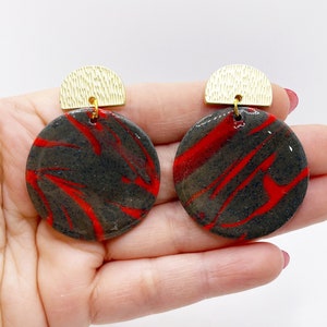 Puede incluir: Pendientes hechos a mano con un diseño veteado gris oscuro y vetas rojas. Los colgantes circulares tienen detalles dorados en forma de media luna. Los pendientes se sostienen en una mano sobre un fondo blanco.