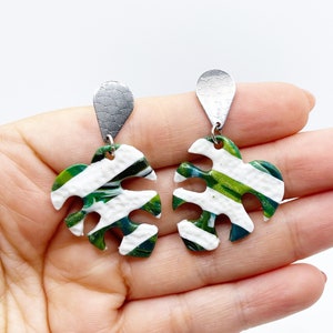 Peut inclure: Une paire de boucles d'oreilles en forme de feuille. Chaque boucle d'oreille présente un fond blanc avec des rayures vertes et vert foncé. Les boucles d'oreilles sont maintenues par une pièce supérieure argentée et texturée. Thème tropical.