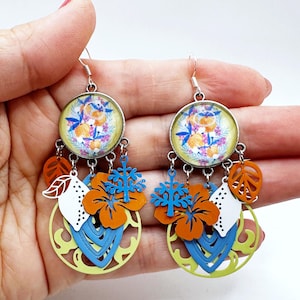 Puede incluir: Un par de pendientes colgantes coloridos. Cada pendiente presenta una parte superior redonda con estampado floral, con formas recortadas en naranja, azul y blanco, que incluyen hojas, flores y un diseño de copo de nieve. Los pendientes son sostenidos por una mano.