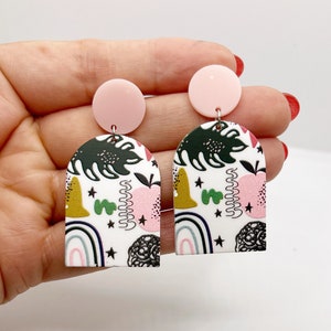 Può includere: Un paio di orecchini dal design astratto. Ogni orecchino presenta un perno circolare rosa, collegato a un ciondolo bianco a forma di arco. I ciondoli sono decorati con un mix di foglie verdi, mele rosa, forme gialle, arcobaleni e scarabocchi neri.