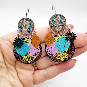 Puede incluir: Pendientes colgantes con un diseño vibrante. Cada pendiente presenta una parte superior circular con un estampado de leopardo colorido, conectado a un círculo de filigrana negro. Tienen detalles de hojas y flores naranjas, azules y moradas.