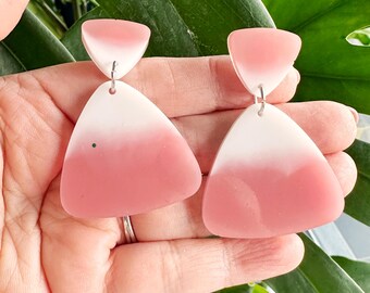 May include: A pair of pink and white geometric earrings. Each earring features a small, white triangle at the top, connected to a larger, downward-pointing triangle that fades from white to pink. The earrings are held in a hand.
