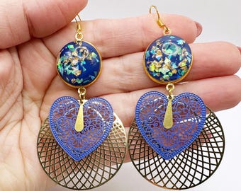 May include: A pair of dangling earrings with gold-colored hooks. Each earring features a dark blue circular top with iridescent accents, a blue heart-shaped center, and a gold-colored, openwork, circular base. The earrings are displayed against a white background.