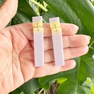 May include: A pair of rectangular earrings with a gradient design. The top section is yellow, fading into a light purple. The earrings are connected to silver hooks. The background features green leaves.