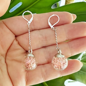 以下が含まれることがあります： 乾燥したピンクの花が詰められた透明なガラスのオーブが特徴のぶら下がりピアス。 ピアスはシルバーカラーの金属アクセントとレバーバッククロージャーを備えています。 緑の葉の背景に表示されています。