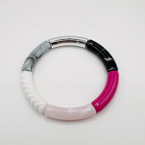 May include: A colorful bracelet featuring a silver metal tube and segments of black and white marbled, white twisted, light pink, and hot pink beads. The bracelet is circular and sits on a white background.