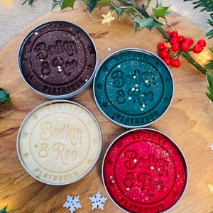 May include: Four tins of sensory playdough in shades of brown, teal, cream, and red. Each tin features the embossed words "Bodkin & Roo" and "Playdough", with gold glitter. The tins are displayed on a wooden board, decorated with holly and snowflake ornaments, creating a festive theme.