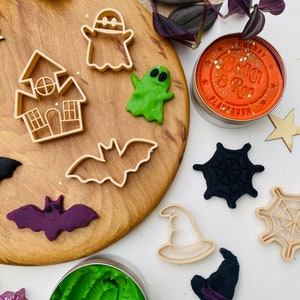 May include: A collection of beige plastic cookie cutters in shapes of a ghost, a haunted house, a bat, a witch hat, and a spider web, alongside orange and green playdough in metal tins, and moulded playdough shapes of a ghost, bat, and spider web.