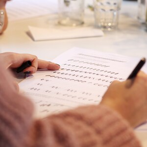 Op de afbeelding: Een persoon oefent kalligrafie op een wit vel papier met een zwarte pen. Het papier heeft lijnen met oefenstreken en lettervormen. De persoon draagt een roze trui en wijst met de wijsvinger naar het papier.