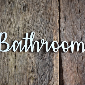 May include: White wooden sign with the word "Bathroom" written in cursive script. The sign is on a rustic wooden background.