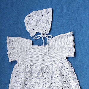 Può includere: Un abito da neonato bianco all'uncinetto con un berretto e scarpine abbinati. L'abito ha un orlo a balze e un laccio in vita.