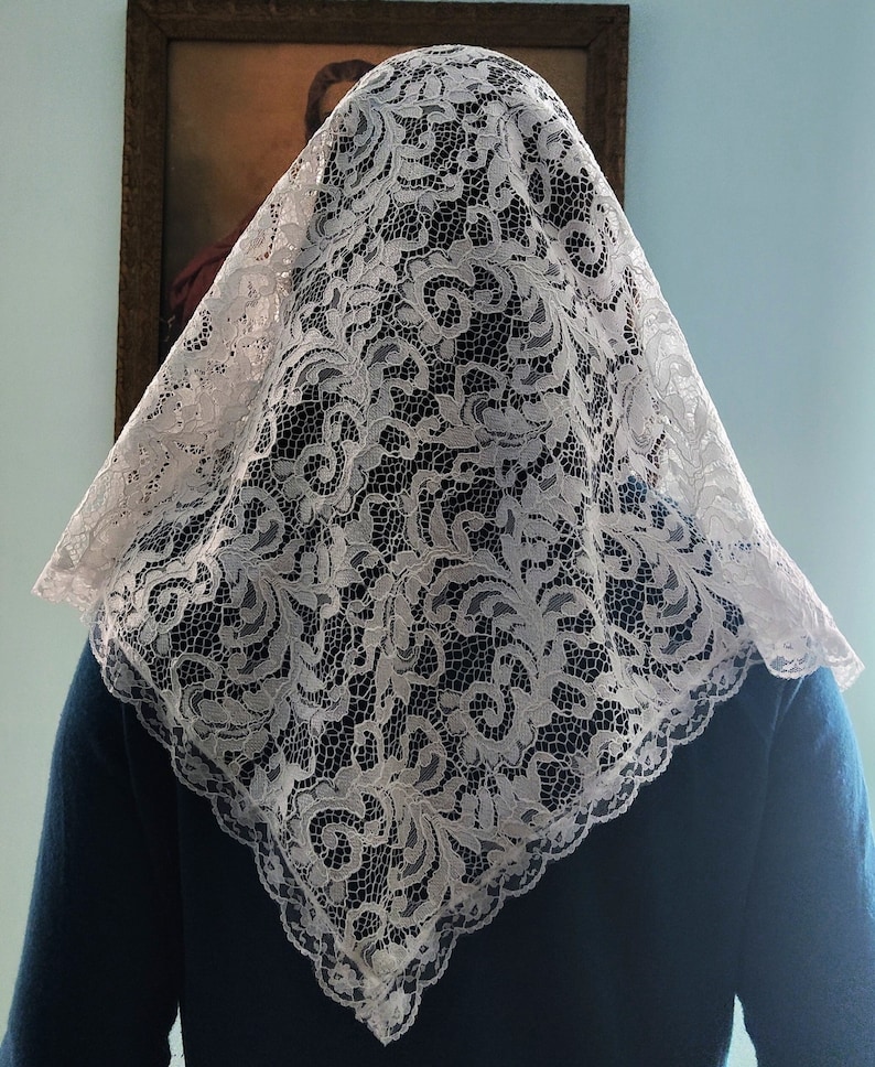 May include: A white lace veil with an elaborate floral pattern, covering the head and shoulders. The square-shaped veil has a scalloped edge. The person is wearing a dark blue top. A framed painting is in the background.