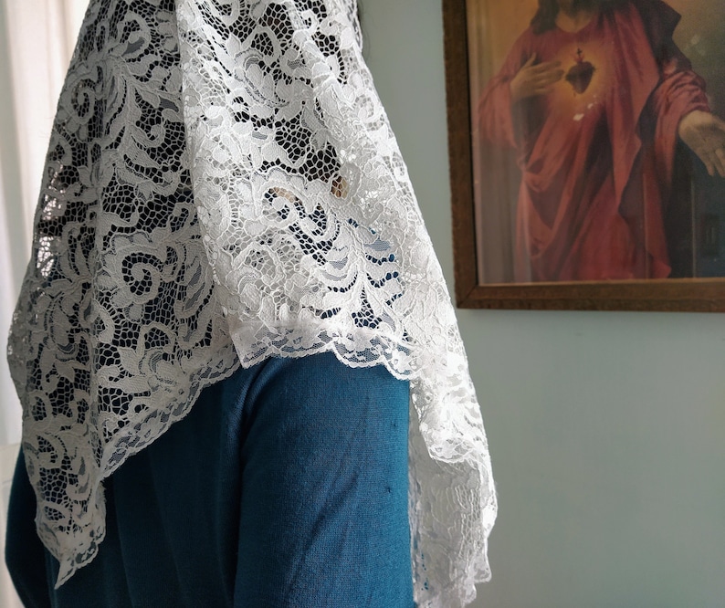 May include: A white lace veil with a detailed floral design. The veil is worn over a teal blue sweater. A framed religious image is visible in the background.